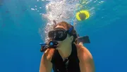 A person is snorkeling underwater, exhaling bubbles near a floating, yellow snorkel tube.