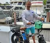 A man in casual clothing is smiling while standing with his hand on the handlebar of a fat tire electric bicycle at a marina with boats docked in the background