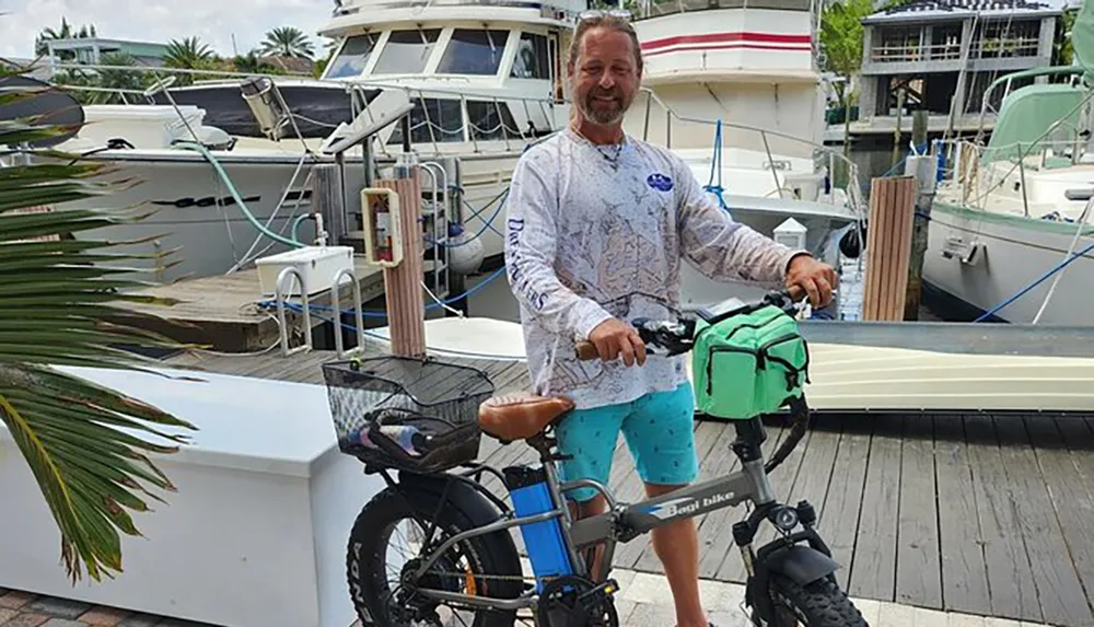 A man in casual clothing is smiling while standing with his hand on the handlebar of a fat tire electric bicycle at a marina with boats docked in the background