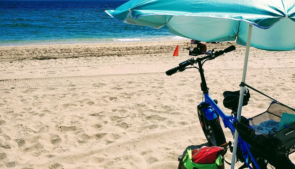 A blue bicycle with a trailer is parked on a sandy beach under a light blue shade umbrella with a view of the turquoise sea