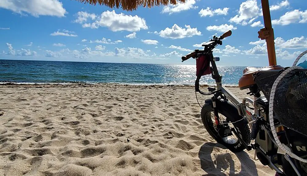 An electric bicycle is parked on a sandy beach with a view of the ocean under a sunny sky