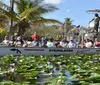 A group of passengers is seated on an airboat named Everglades Safari gliding through a waterway lined with lily pads with a guide standing at the helm on a sunny day