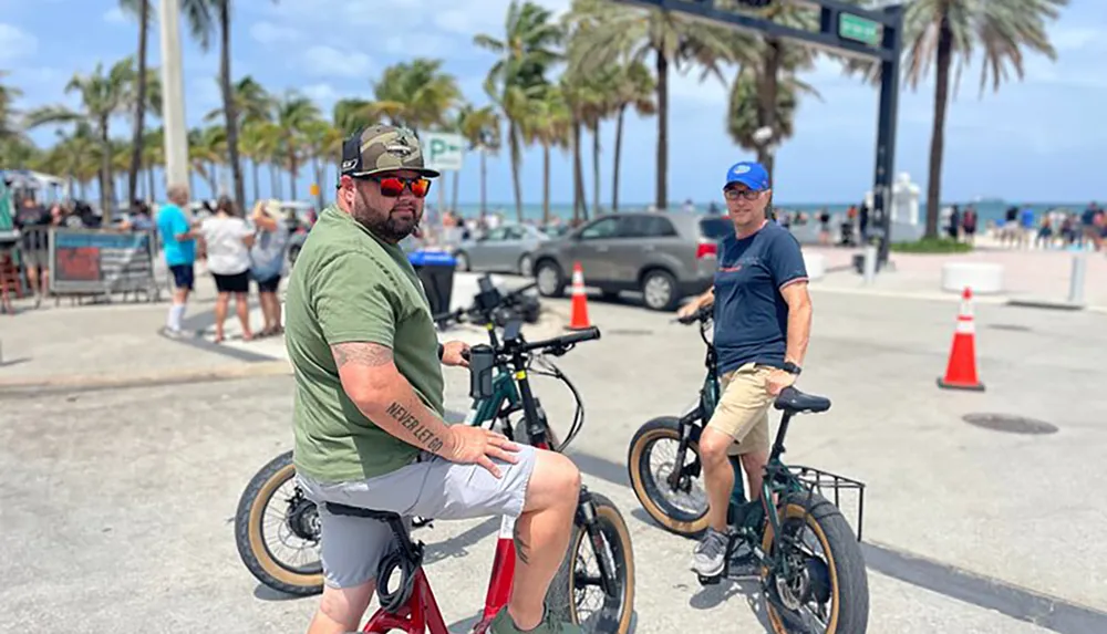 Two men are leisurely riding their bicycles by a beachside promenade lined with palm trees on a sunny day