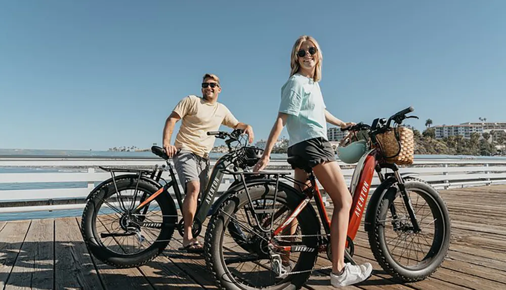 Two people are smiling and standing next to their bicycles on a sunny pier with a clear blue sky and the ocean in the background