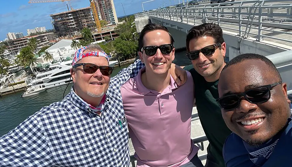 Four smiling men are taking a group selfie on a sunny day with a marina featuring boats and construction in the background