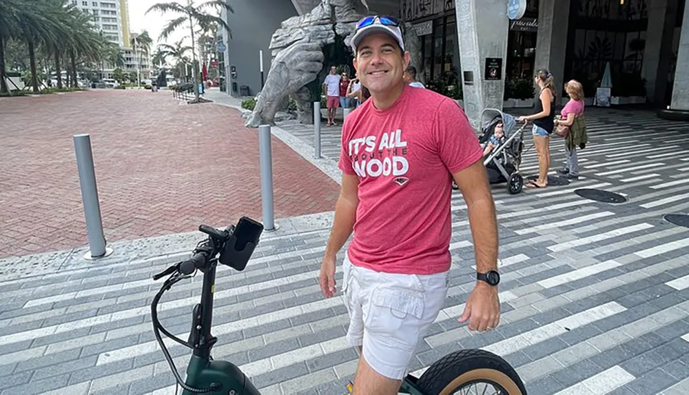 A smiling person stands on a patterned pavement near a scooter with tropical trees and a statue in the background