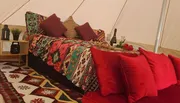 The image shows a cozy, bohemian-style glamping setup with a colorful patterned bedspread, matching carpet, red pillows, and romantic touches like a bottle of wine and red roses.
