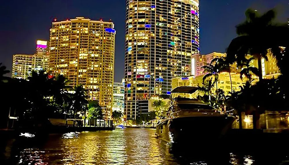 The image showcases a nighttime scene of a vibrant waterfront cityscape with illuminated high-rise buildings and luxury yachts docked along the river