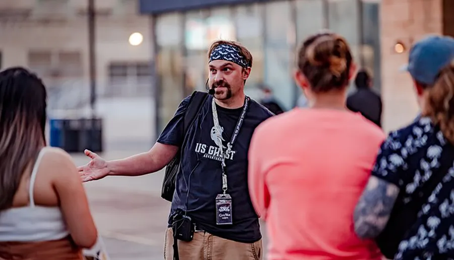 A man wearing a headband, microphone headset, and a black t-shirt is gesturing while speaking to a group of people on the street.