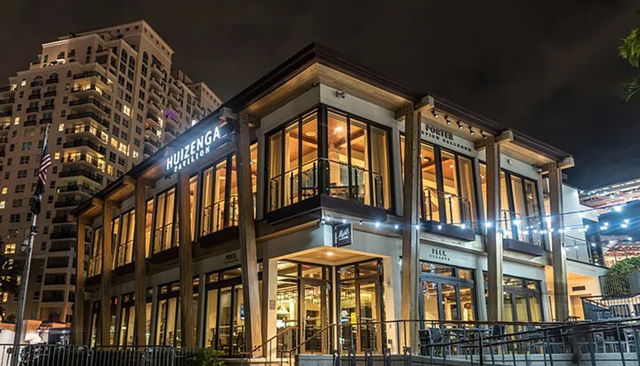 The image shows a two-story building with warm interior lighting and a prominent sign HUIZENGA PAVILION at night, set against a backdrop of a high-rise residential building.