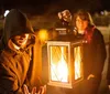 A hooded figure holds a lantern casting a warm glow as people gather closely around in a nighttime outdoor setting