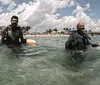 Two scuba divers are standing in shallow sea water near the shore equipped with diving gear and smiling with a sunny beach and clouds in the background