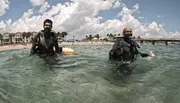 Two scuba divers are standing in shallow sea water near the shore, equipped with diving gear and smiling, with a sunny beach and clouds in the background.
