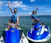 Two individuals are joyfully raising their arms while standing on stationary jet skis in a sunny open water setting with a coastline in the distance