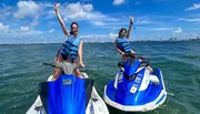 Two individuals are joyfully raising their arms while standing on stationary jet skis in a sunny, open water setting with a coastline in the distance.