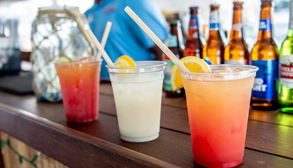 Three colorful cocktails with eco-friendly straws are in the foreground on a bar counter with a blurred background of beer bottles and a person in blue