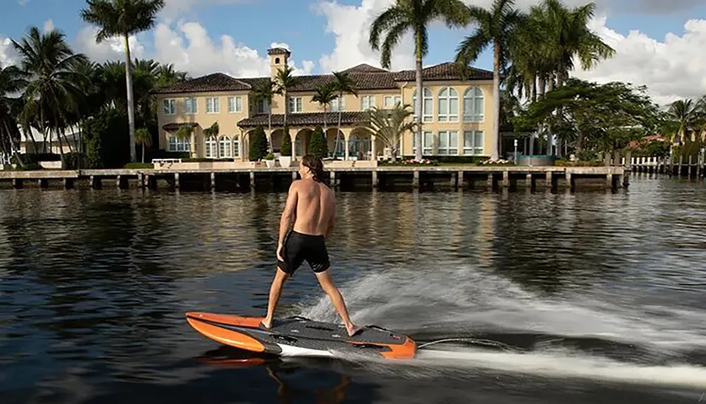 A person is riding an electric surfboard on the water near a large luxurious waterfront house surrounded by palm trees