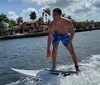 A person is wake surfing on a wave created by a boat on a sunny day with palm trees and houses in the background