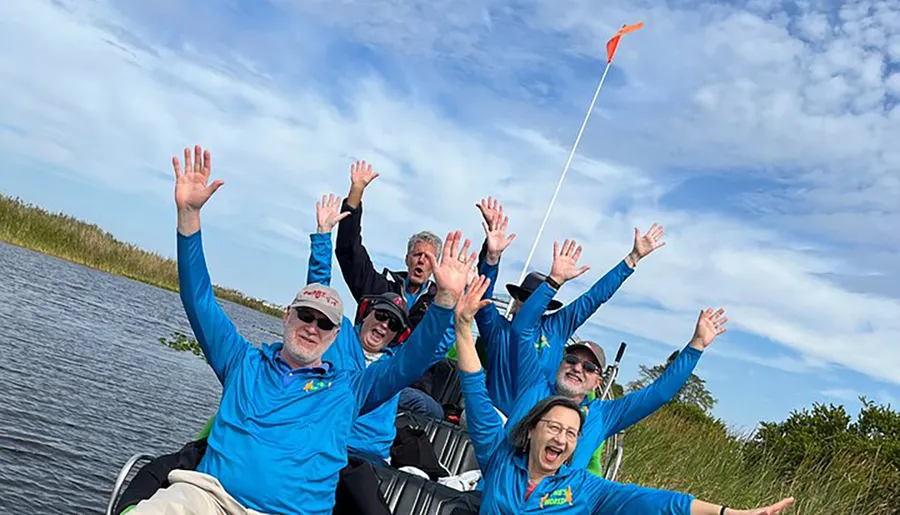 A group of people in matching blue shirts are joyfully raising their hands while on a boat ride under a clear blue sky.