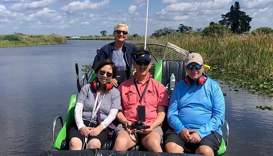 Four individuals are seated on an airboat in a waterway, likely in a marsh or wetland environment, with protective ear muffs on and smiling for the camera.