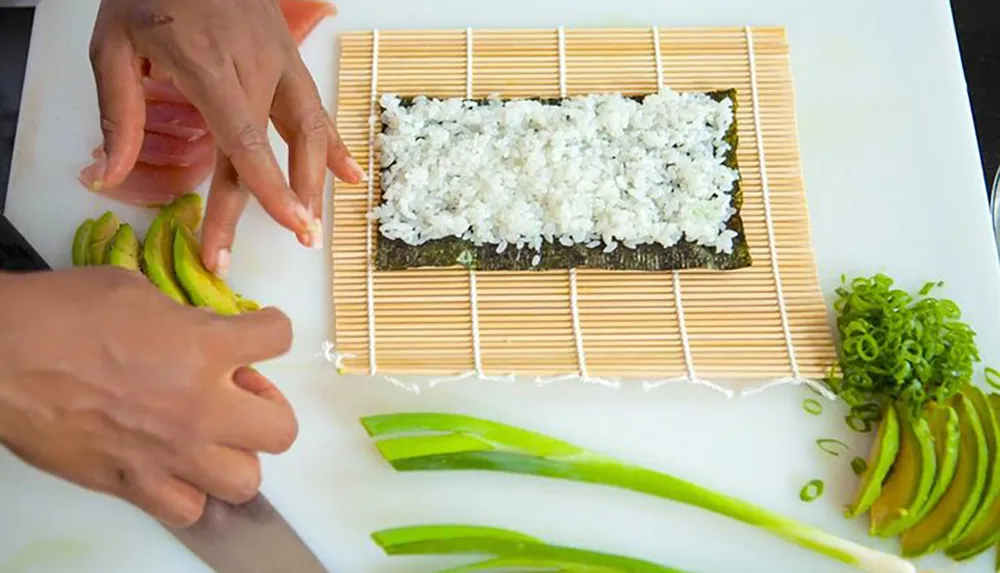 A person is preparing sushi arranging sliced avocado and rice on a sheet of nori placed on a bamboo rolling mat