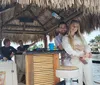 A couple is smiling and embracing near a bamboo bar with a thatch roof on a boat with two other individuals in conversation behind them