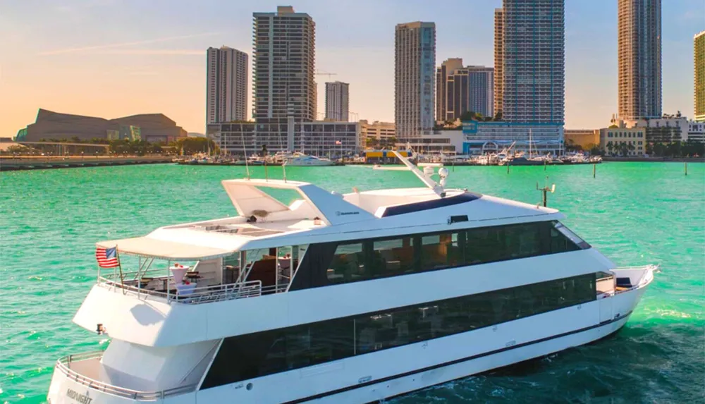 A luxury yacht is cruising in clear turquoise waters near a city skyline with modern high-rise buildings under a bright blue sky