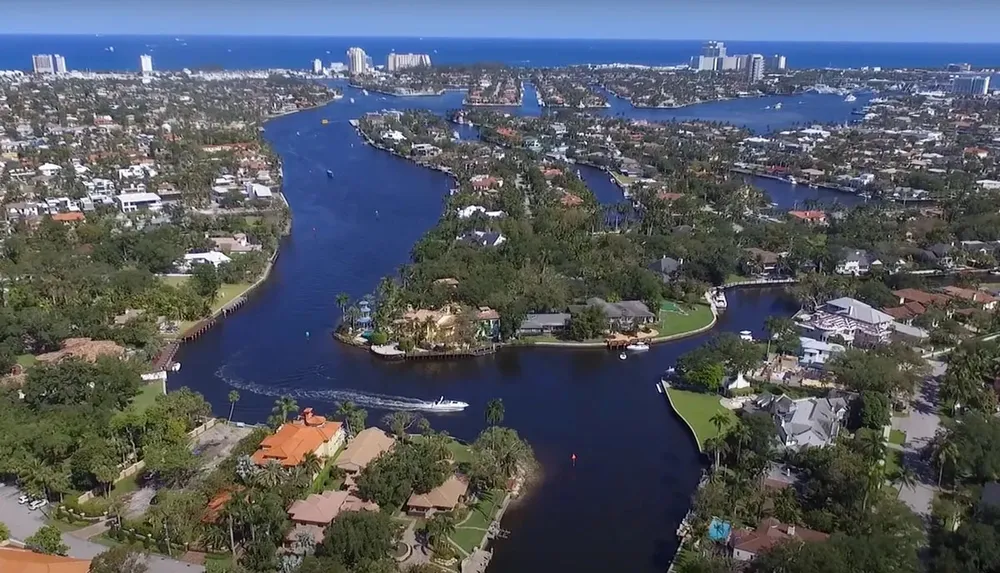 This is an aerial view of a winding river with boats surrounded by luxury homes and lush greenery leading towards a skyline by the coast