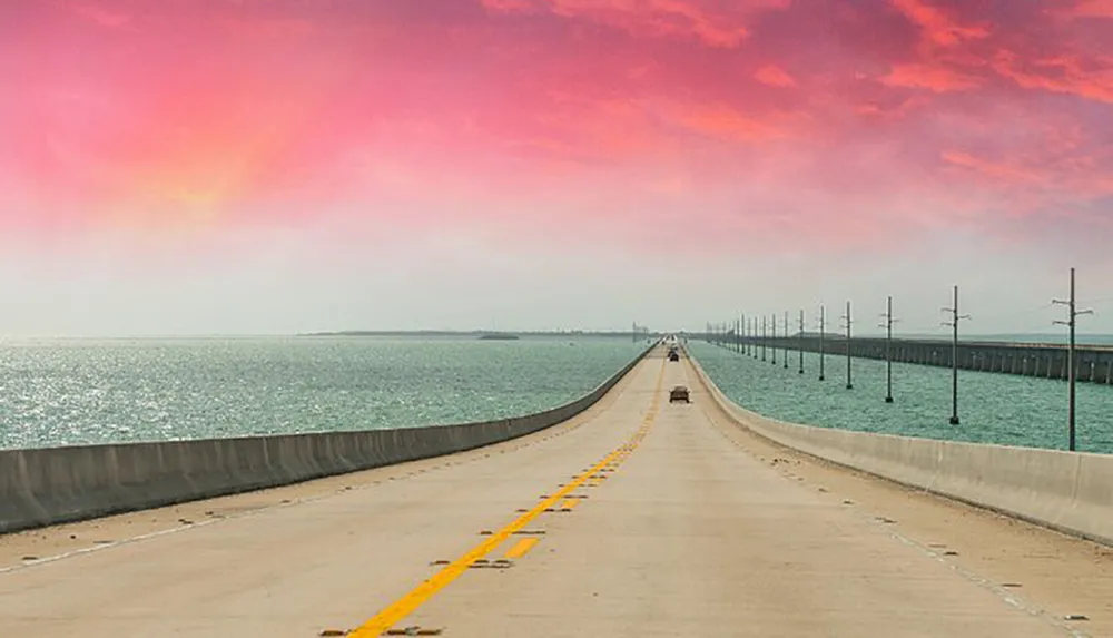 The image shows a long bridge stretching across a body of water under a sky painted with vibrant shades of pink and orange from the setting or rising sun