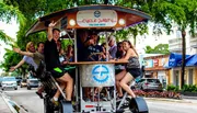A group of happy people are enjoying a ride on a multi-passenger, pedal-powered vehicle in an urban setting.