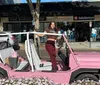 A person is posing with a smile on a parked pink open-topped vehicle on a sunny day outside of a shopping area
