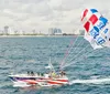 A parasailer is flying above the ocean towed by a speedboat with a coastal city skyline in the background