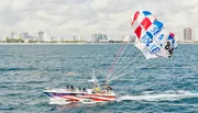 A parasailer is flying above the ocean, towed by a speedboat, with a coastal city skyline in the background.