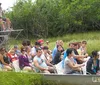 A group of people are seated in a flat-bottomed airboat on a tour with the guide standing on an elevated platform at the back