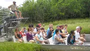 A group of people are seated in a flat-bottomed airboat on a tour, with the guide standing on an elevated platform at the back.