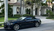 A black convertible car is parked outside a luxurious two-story house with palm trees and a clay tile roof.