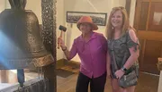 Two smiling women pose beside a large bell, one holding a mallet likely to ring the bell.
