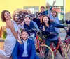A group of joyous people including a bride and groom are posing with bicycles on a sunny street exhibiting expressions of celebration and happiness