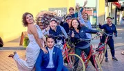 A group of joyous people, including a bride and groom, are posing with bicycles on a sunny street, exhibiting expressions of celebration and happiness.