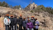 A group of people wearing orange helmets stands smiling in front of a stone building resembling a chapel atop a rocky hillside.