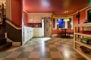 The image shows a colorful kitchen interior with a checkered floor, warm lighting, and a small dining area visible through an archway.