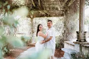 A couple is posing affectionately under a vine-covered pergola in a lush garden setting.