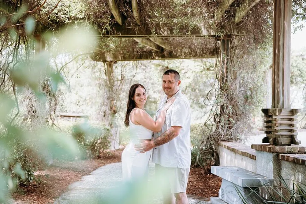 A couple is posing affectionately under a vine-covered pergola in a lush garden setting