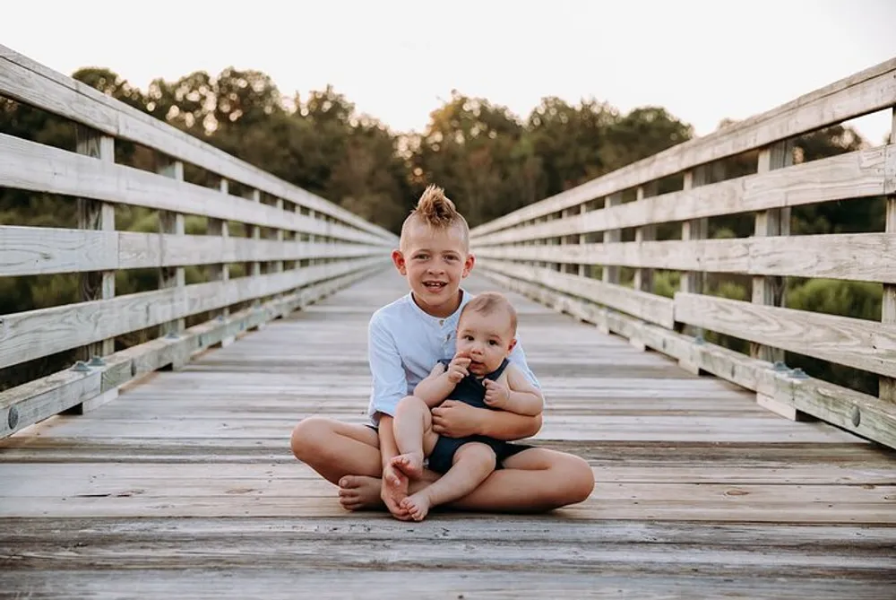 The image captures a smiling older child with his arm around a younger child sitting together on a wooden bridge during what appears to be late afternoon