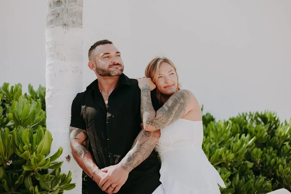 A tattooed couple is embracing while smiling near a palm tree in a sunny outdoor setting