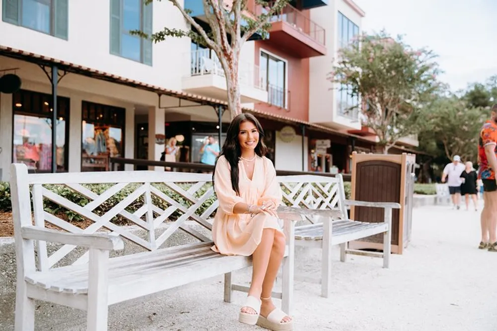 A smiling person is seated comfortably on a white bench in a pleasant outdoor shopping area