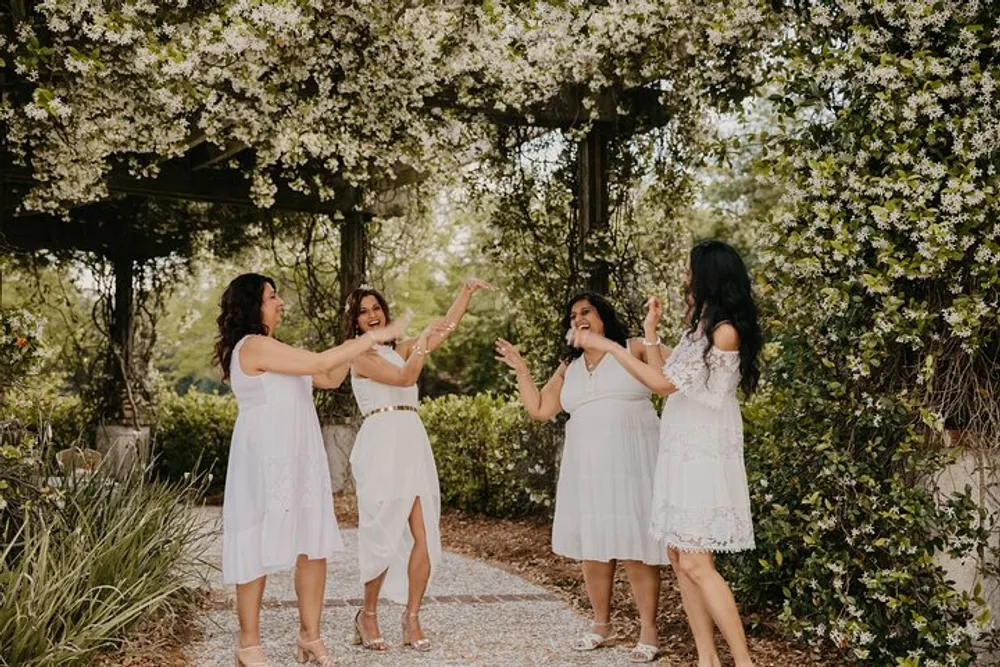 Four women in white dresses are joyfully interacting with each other beneath a canopy of blooming flowers in a garden setting