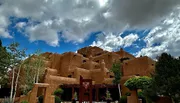 The image depicts an adobe-style building with a terraced structure under a blue sky dotted with fluffy clouds.