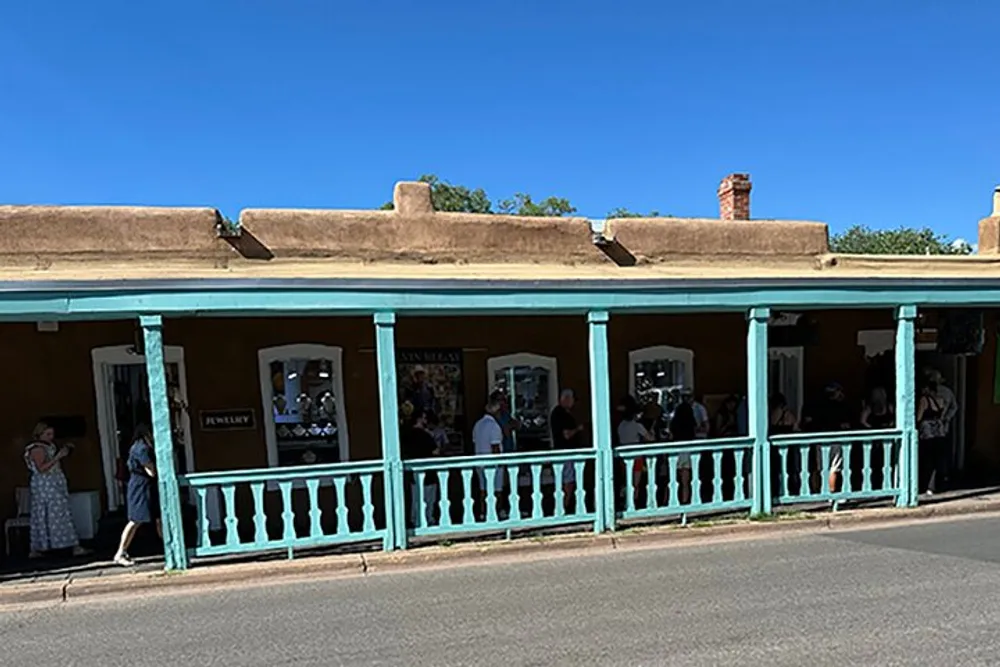 The image portrays a sunny day where people are walking by and others are inside an adobe-style building with a turquoise porch railing