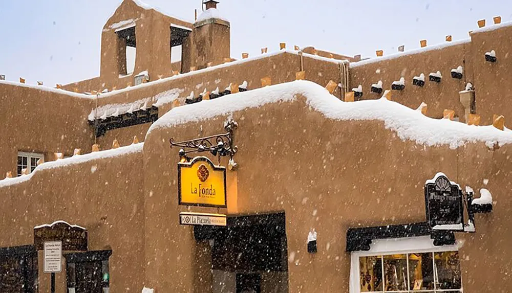 A traditional pueblo-style building is covered in snow with the La Fonda sign visible during a heavy snowfall